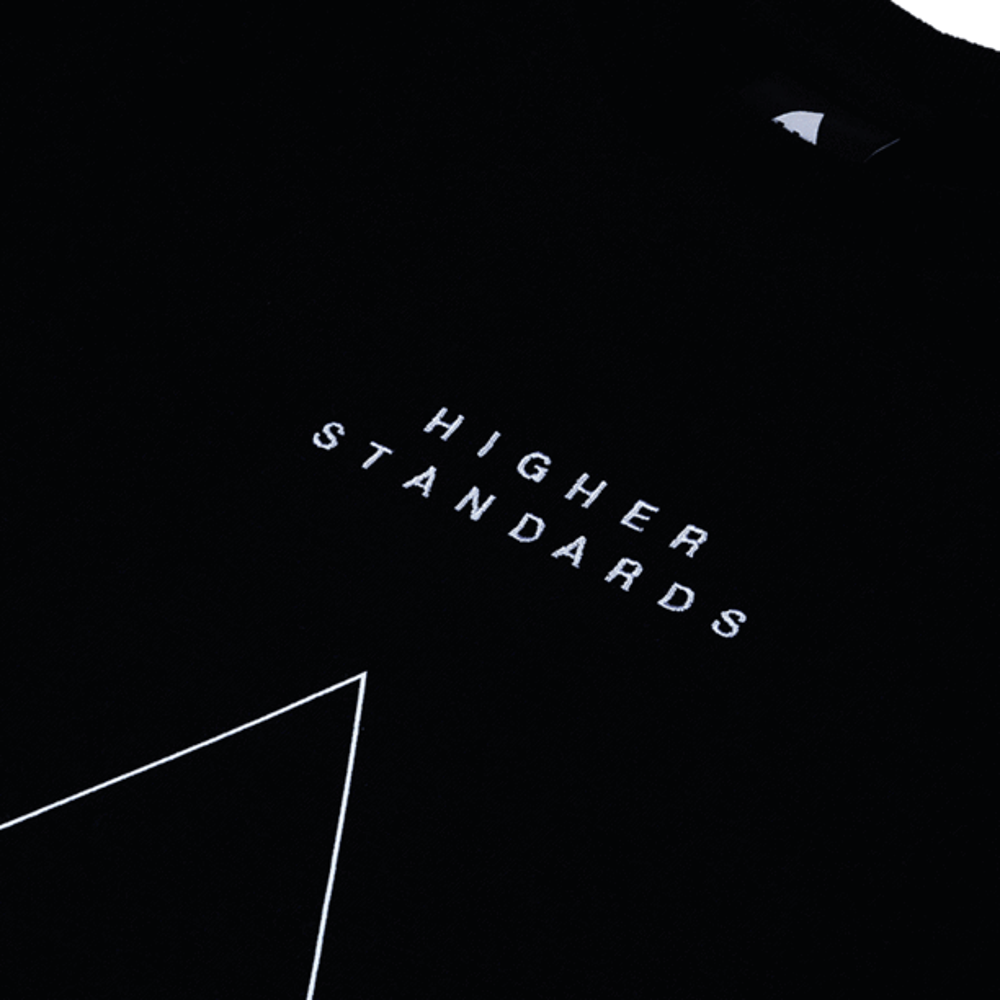 Close-up detail shot focusing on the high-quality white embroidery of the 'HIGHER STANDARDS' text and triangle design on black cotton fabric.