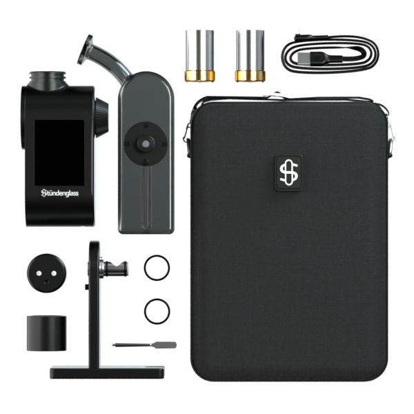 A comprehensive, top-down studio layout shot displaying the entire Stündenglass Modül + Dok Deluxe Travel Set with Gray Glass. The open hardshell carrying case sits at the top, flanked by the assembled Dok station with its smoky gray glass globe, the digital Modül battery unit, both dry material and quartz concentrate heating tanks, the magnetic connection rings, a metal loading tool, and the USB-C fast-charging cable spread neatly on a clean background.
