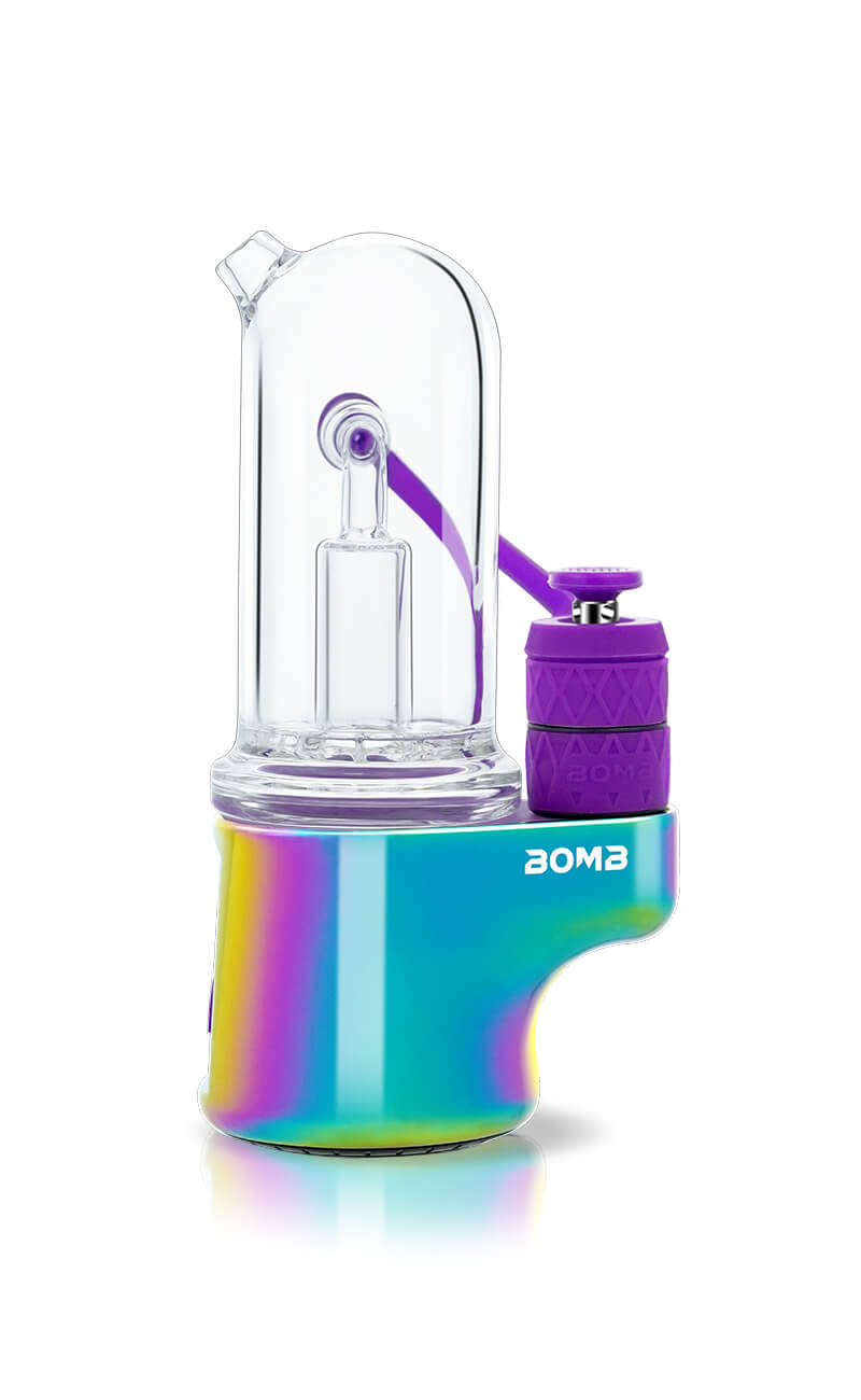 A side profile of the Bomb Erig Aerix in Rainbow, showcasing the "Bomb" logo on the multi-color PVD base. This shot highlights the detachable purple airpath tube and the matching purple carb cap that connect to the glass bubbler.