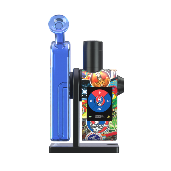 A crisp, front-facing studio product shot showcasing the Grateful Dead x Stündenglass Modül + Dok Legacy Patchwork edition. The shot highlights the premium handheld Dok station with its clear borosilicate glass globe on top, the sleek 2.4-inch digital color display screen active on the front, and the intricate, multicolored montage of official licensed Grateful Dead artwork decorating the entire lower chassis frame.