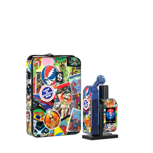 A front-angle studio product shot displaying the assembled Grateful Dead x Stündenglass Modül + Dok Legacy Patchwork device standing next to its matching hardshell travel case. The handheld glass e-rig station showcases vibrant, colorful Grateful Dead patchwork graphics wrapped around its base, perfectly complementing the heavy-duty zippered carrying case featuring identical iconic band imagery.