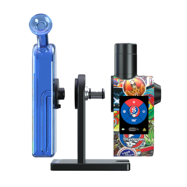 A detailed studio product shot demonstrating the assembly of the Grateful Dead x Stündenglass Modül + Dok Legacy Patchwork set. The hands-free display illustrates the Modül smart heating engine and the clear borosilicate glass globe attachment being aligned and installed onto the magnetic connection ports of the custom patchwork-wrapped Dok station base.
