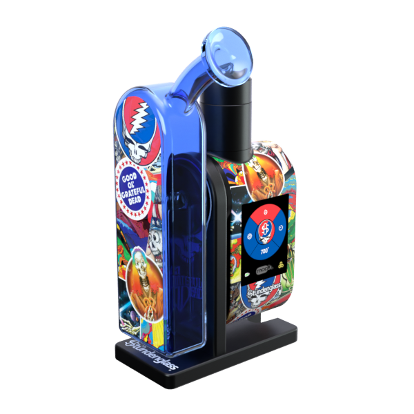 A clean front angle profile studio shot of the assembled Grateful Dead x Stündenglass Modül + Dok Legacy Patchwork device. This view showcases the ergonomic curve of the handheld docking handle, highlighting the seamless continuation of the intricate, multicolored licensed Grateful Dead patchwork graphics extending around the side of the chassis beneath the clear glass globe.