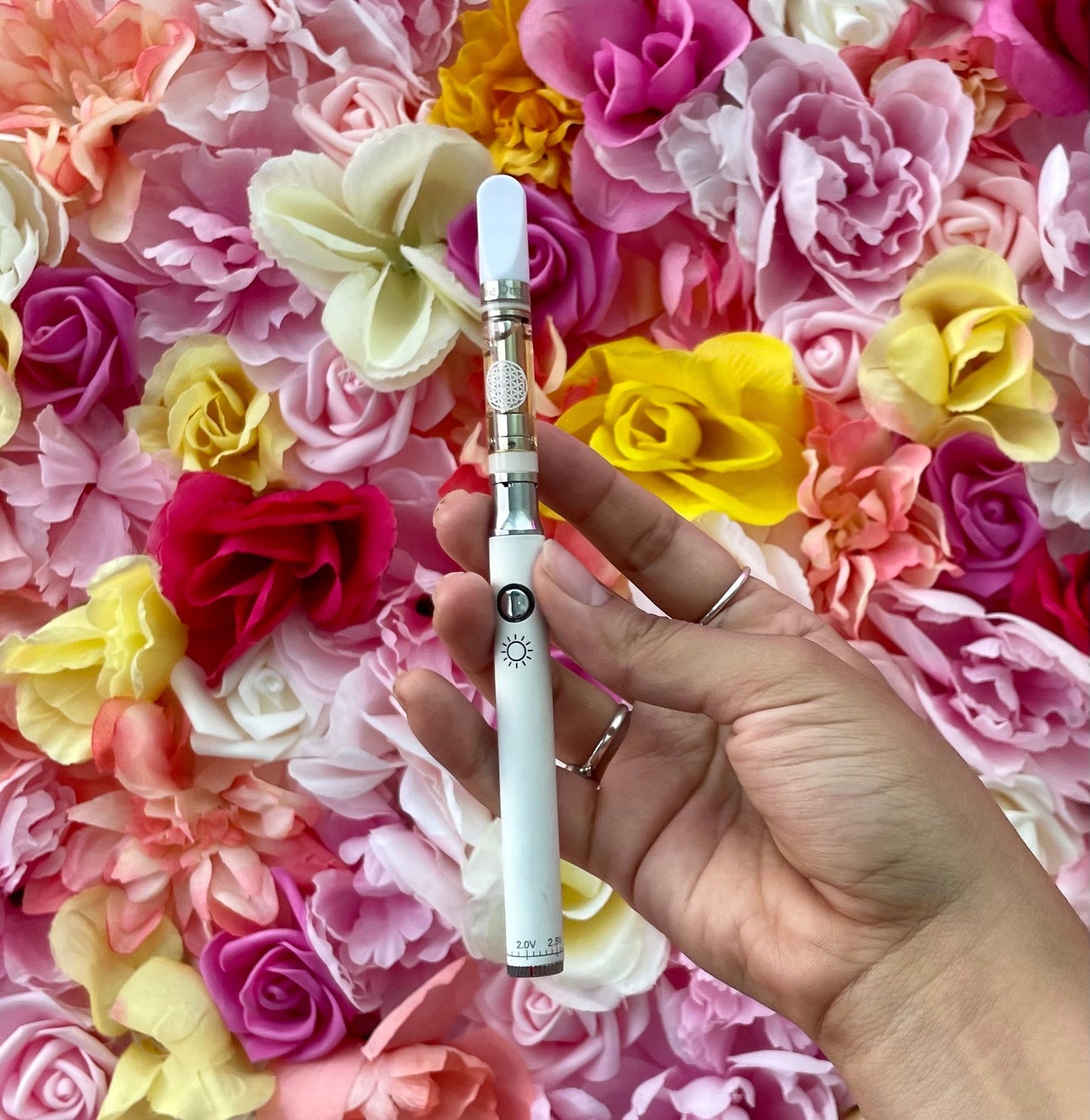 A lifestyle view of the Jane West Day Battery being held in a hand with a 510 cartridge installed. The background features a soft floral arrangement, emphasizing the device's sophisticated and natural design.