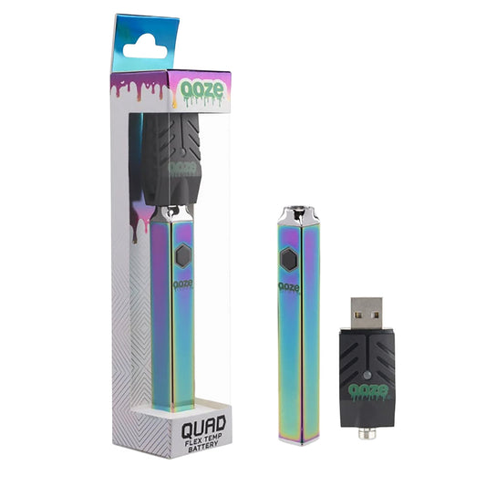 A crisp studio product shot displaying the Ooze Quad Flex Temp square 510 battery in the vibrant Rainbow colorway. The metallic, multi-colored neo-chrome pen sits vertically next to its transparent plastic retail pegboard packaging, with the included black 510-threaded USB smart charger laid out neatly in the foreground.