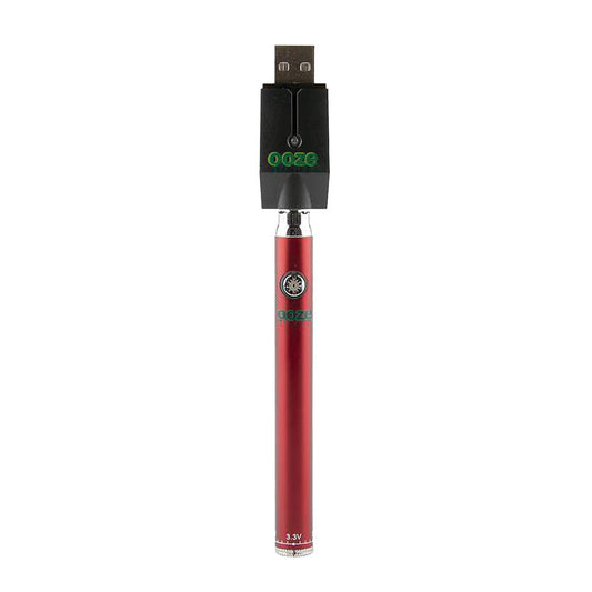 The Ruby Red Ooze Slim Twist Pen shown screwed into the included Ooze Smart USB Charger. This shot demonstrates the charging setup, highlighting the deep red metallic finish of the battery and the red/green LED indicator light on the charger.