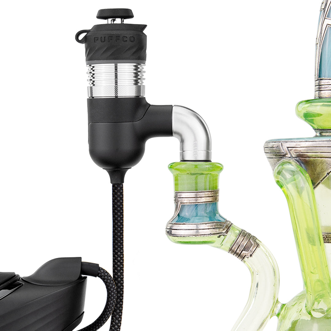 A dynamic, semi-close-up studio product shot showing the Puffco Peak Pro Link seated directly into the joint of a clear glass water pipe rig. The photo illustrates how the silicone adapter joint forms a snug seal, bridging the 30-inch tether cable from the Peak Pro power unit to the traditional glass piece.