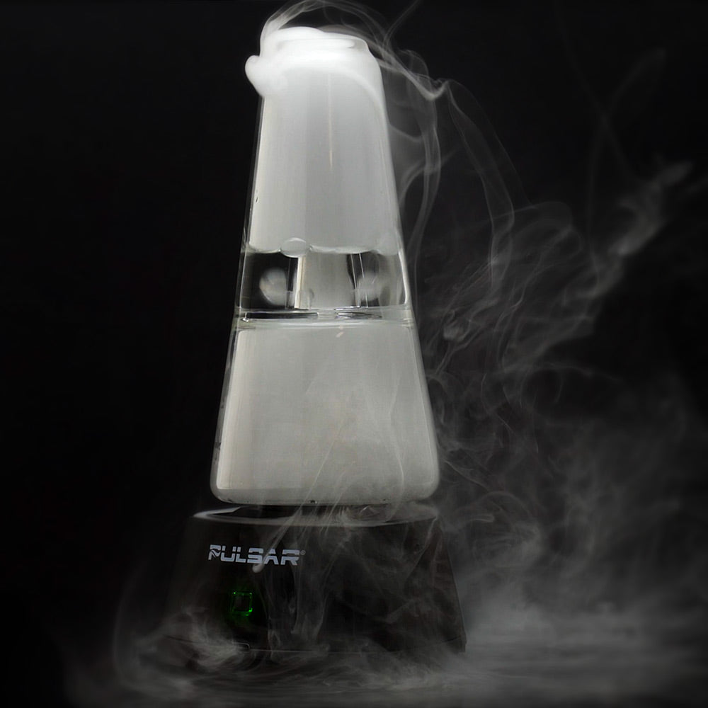 A studio shot of the Pulsar Sipper Bubbler Cup Edition in use, filling with dense white vapor. The glass bubbler is attached to the illuminated black base, showcasing the hands-free auto-fill pump feature as thick vapor bubbles through the water reservoir and pools inside the clear glass chalice.
