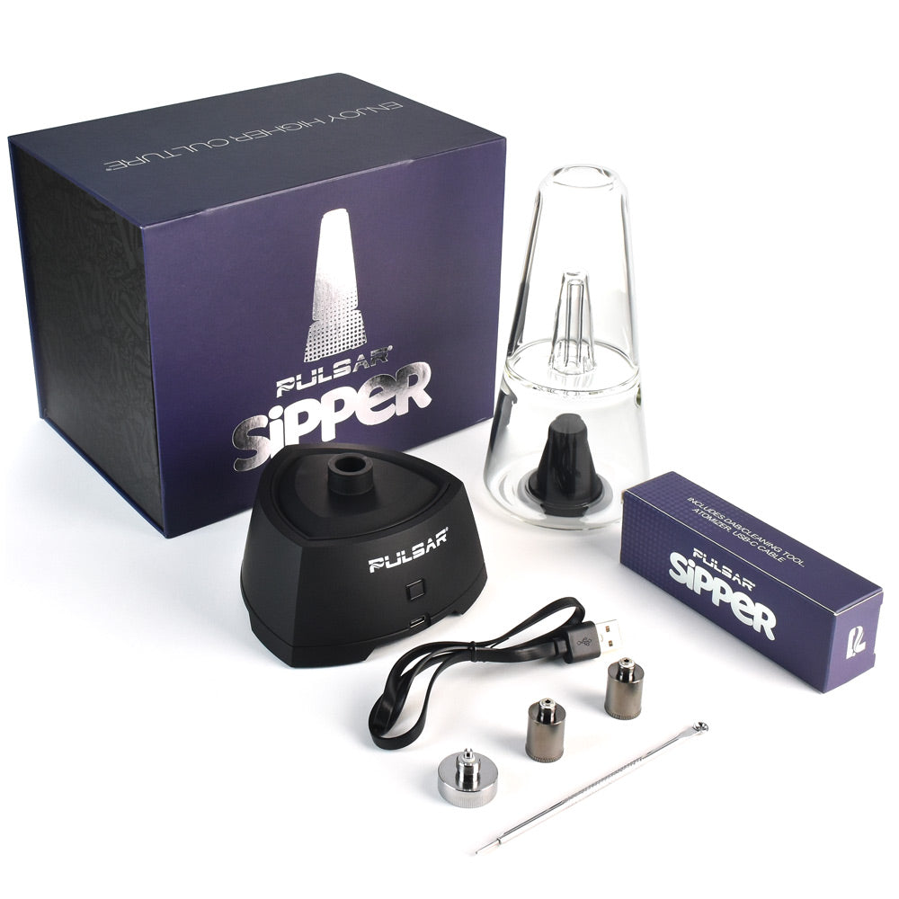 A studio layout showing the Pulsar Sipper Bubbler Cup Edition retail box with the entire kit unpacked and arranged in front. The display includes the sleek matte black base, the clear borosilicate glass bubbler cup, the triple quartz coil concentrate atomizer, a 510-threaded adapter, a protective chamber cover, a stainless steel dab tool, and a USB-C charging cable.