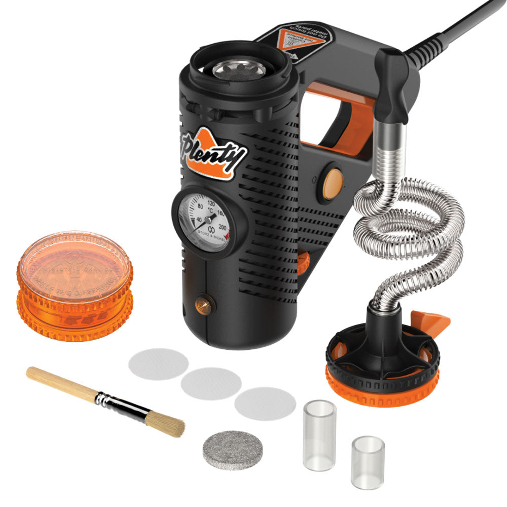 A comprehensive studio layout shot displaying the Storz & Bickel Plenty Vaporizer surrounded by all its included box accessories. The black and orange desktop vaporizer sits in the center, flanked by its stainless steel cooling coil, replacement metal screens, a liquid drip pad, the official orange acrylic herb grinder, a cleaning brush, and the quick-start instruction manual spread neatly on a clean white background.
