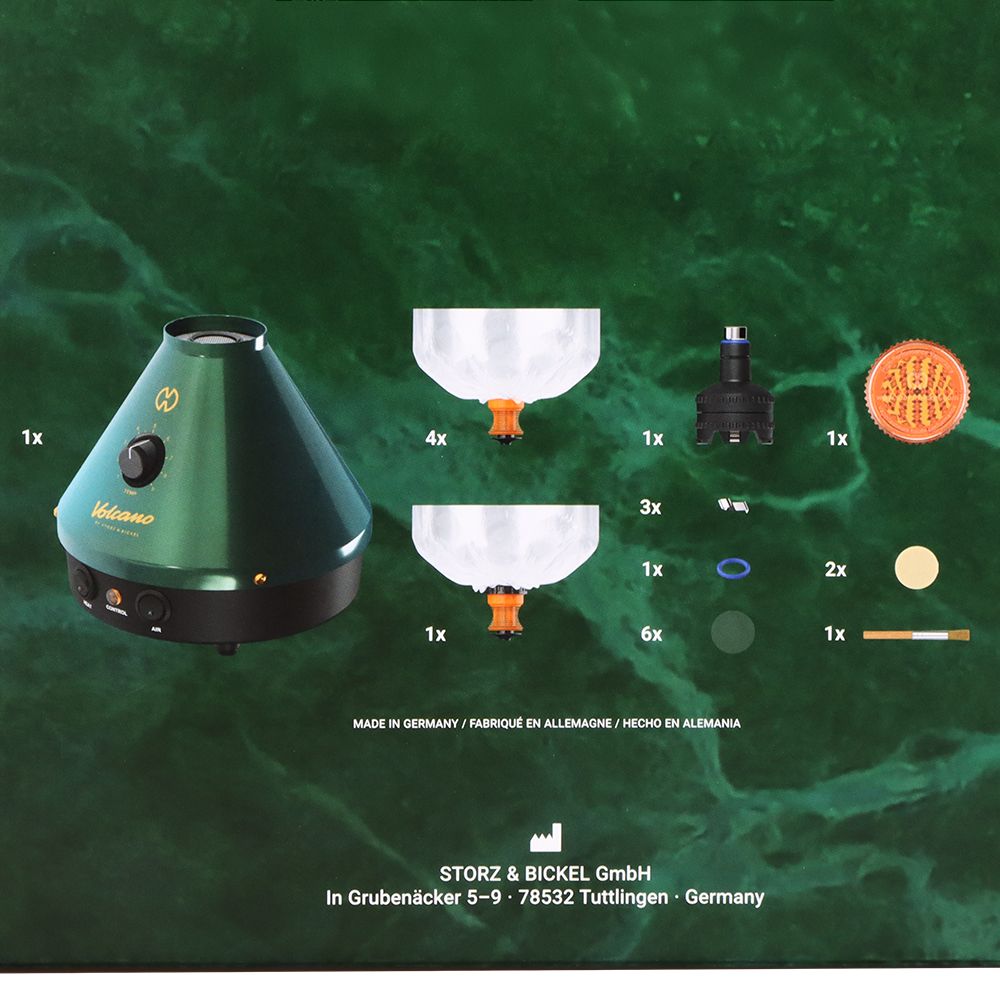 A comprehensive studio product layout displaying the Storz & Bickel Volcano Classic 25-Year Green & Gold Limited Edition next to all included kit accessories. The matte emerald green desktop cone sits on a clean background, surrounded by four Easy Valve balloon bags with mouthpieces, a filling chamber unit, an official orange acrylic herb mill grinder, replacement air filters, a metal screen set, and a cleaning brush.