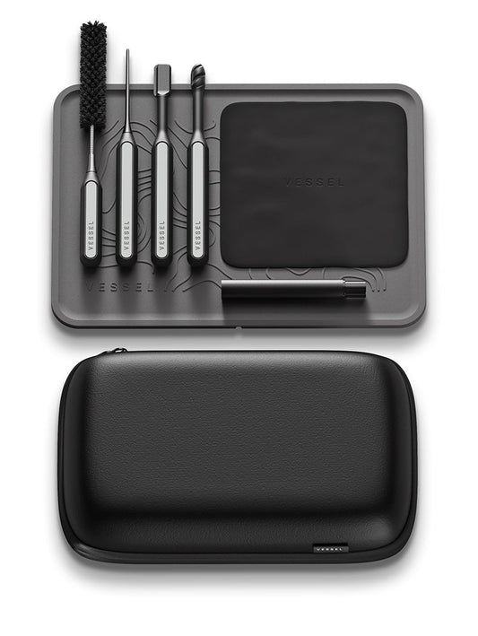A flat-lay view of the Vessel Care Kit showing all seven components. The image features the compact black carry case, the molded EVA interior, four precision cleaning tools (Spiral Swab, Clearing Paddle, Sweeping Brush, Fine Pick), the refillable spray bottle, the microfiber cloth, and the non-slip silicone mat.