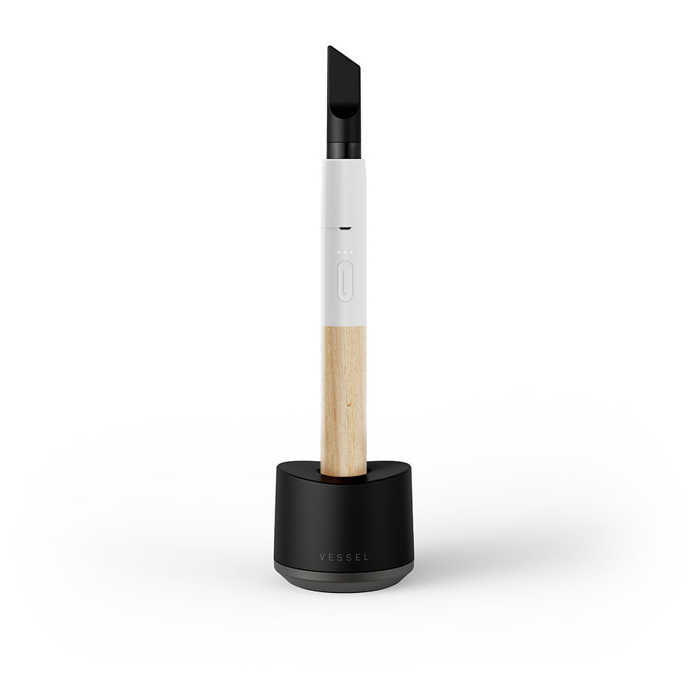 A premium studio product shot showing the Vessel Craftsman vape pen in White and Beechwood standing vertically inside the official Vessel Base Charger. The device, equipped with an installed 510 oil cartridge, sits flush within the heavy-duty black desktop charging dock, showcasing how the magnetic base connections seamlessly align for convenient desktop charging.