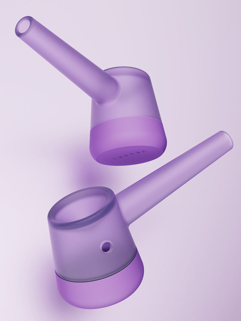Two Vessel Forge Lilac pipes shown together from different angles. One pipe is positioned to show the side profile and ergonomic neck, while the other is angled to showcase the bowl and the unique, hand-sculpted transition between the borosilicate glass and the lilac base.
