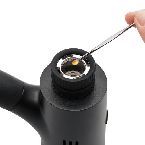 A close-up action shot capturing a stainless steel dab tool loading wax concentrate directly onto the quartz heating chip of the Waxmaid Ares Plus. The perspective focuses on the top-loading atomizer chamber seated above the zinc alloy chassis and clear glass percolator.
